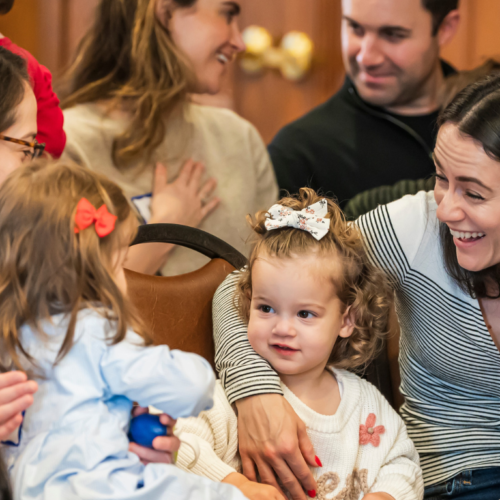 Adults and young children gather with smiles and conversation in a welcoming space at Temple Emanu-El.