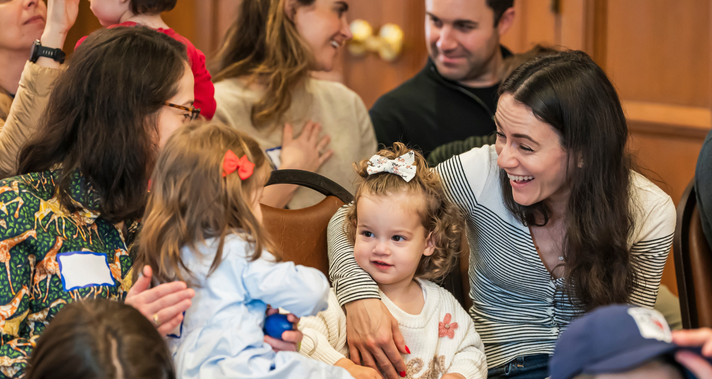 Adults and young children gather with smiles and conversation in a welcoming space at Temple Emanu-El.