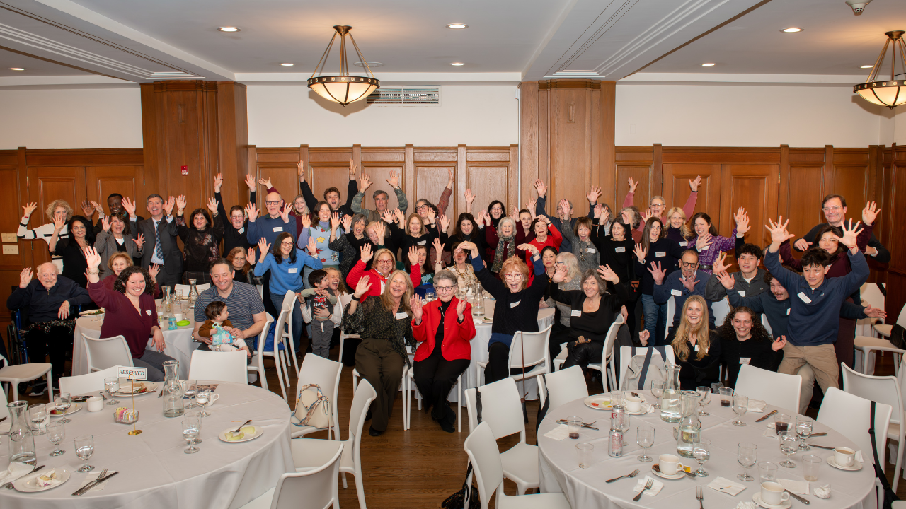 Congregants in Temple Emanu-El’s banquet hall smile and raise their hands, gathered around round tables during a community event.