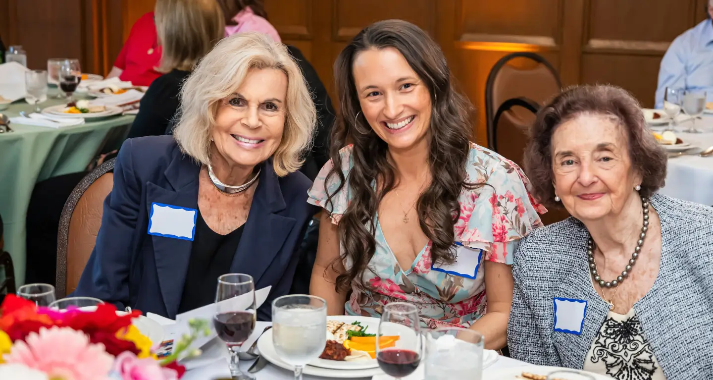 Three women smiling at a formal Temple Emanu-El dinner, wearing name tags, with plates of food and drinks on the table.