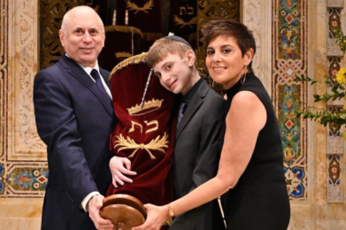 A boy holding a Torah scroll stands between two adults in Temple Emanu-El, all smiling warmly at the camera.