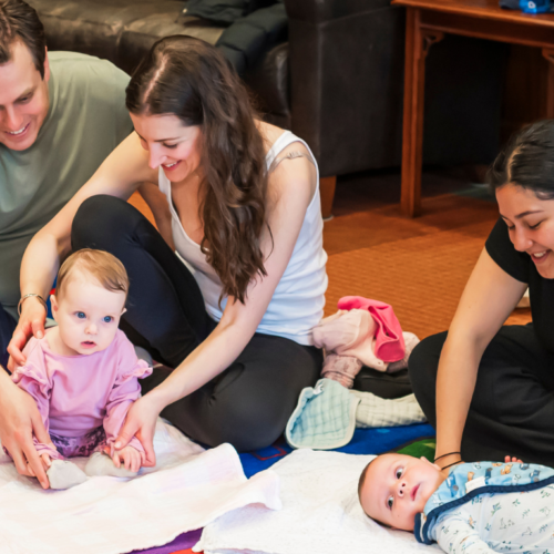 Two adults engage with a baby on a blanket while another adult gently watches over an infant nearby in a welcoming setting.