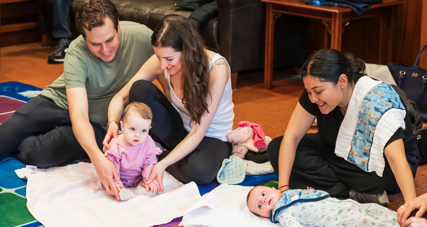 Two adults engage with a baby on a blanket while another adult gently watches over an infant nearby in a welcoming setting.