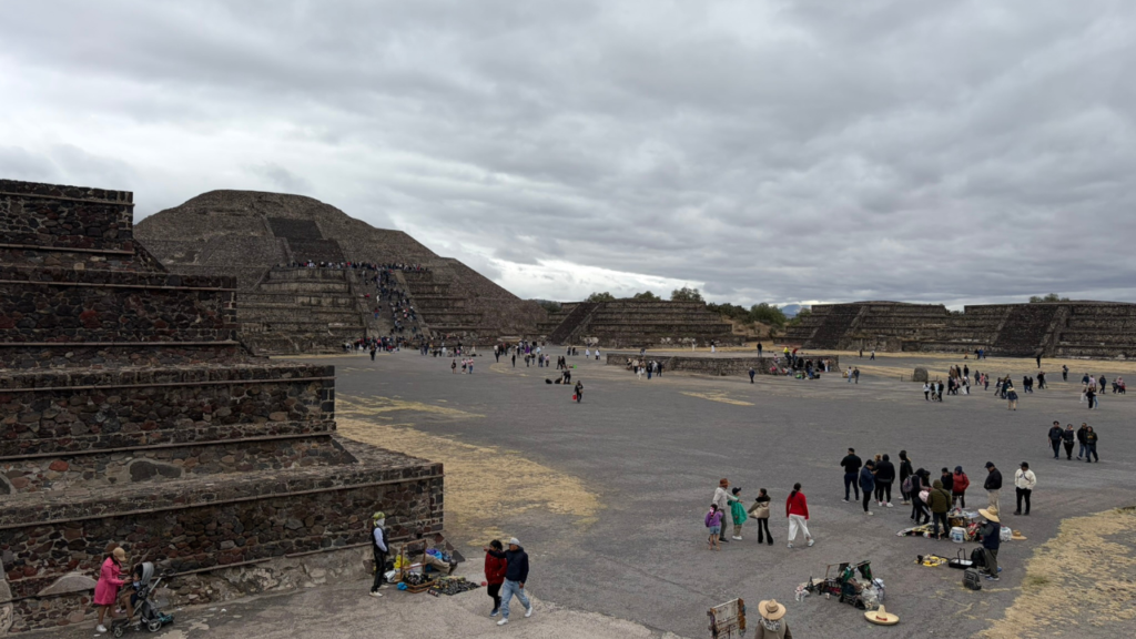 Visitors stroll near the historic pyramids at Teotihuacan, Mexico, beneath a cloudy sky.