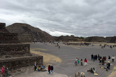 Visitors stroll near the historic pyramids at Teotihuacan, Mexico, beneath a cloudy sky.