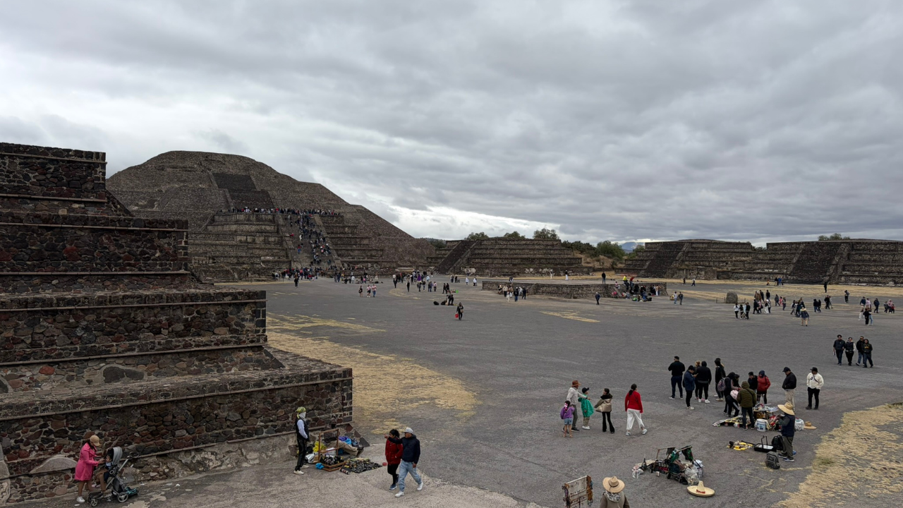 Visitors stroll near the historic pyramids at Teotihuacan, Mexico, beneath a cloudy sky.
