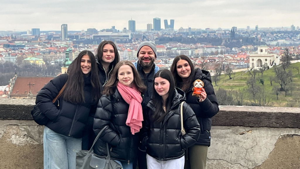 Six congregants in winter jackets smile together with the Prague skyline visible behind them on a cloudy day.