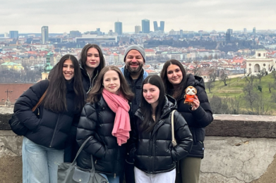 Six congregants in winter jackets smile together with the Prague skyline visible behind them on a cloudy day.