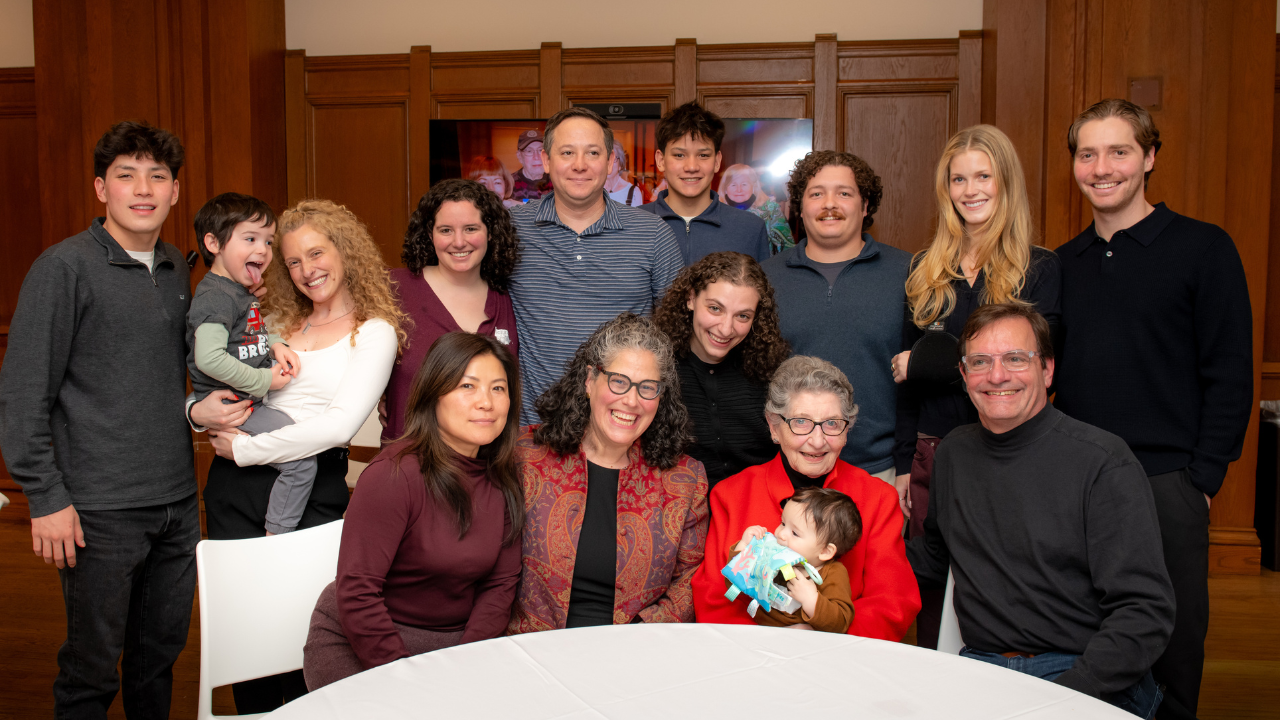 Thirteen people of all ages, including a baby, gather and smile indoors around a white table at Temple Emanu-El in New York City.