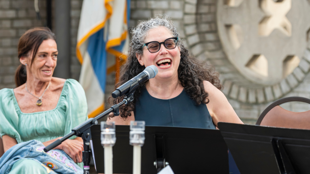 Two women sit outside Temple Emanu-El, one smiling and speaking into a microphone, with music stands before them.