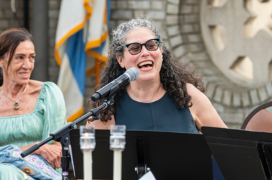 Two women sit outside Temple Emanu-El, one smiling and speaking into a microphone, with music stands before them.