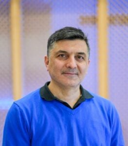 A man with short dark hair and a blue sweater smiles warmly at the camera, with a softly blurred background at Temple Emanu-El.