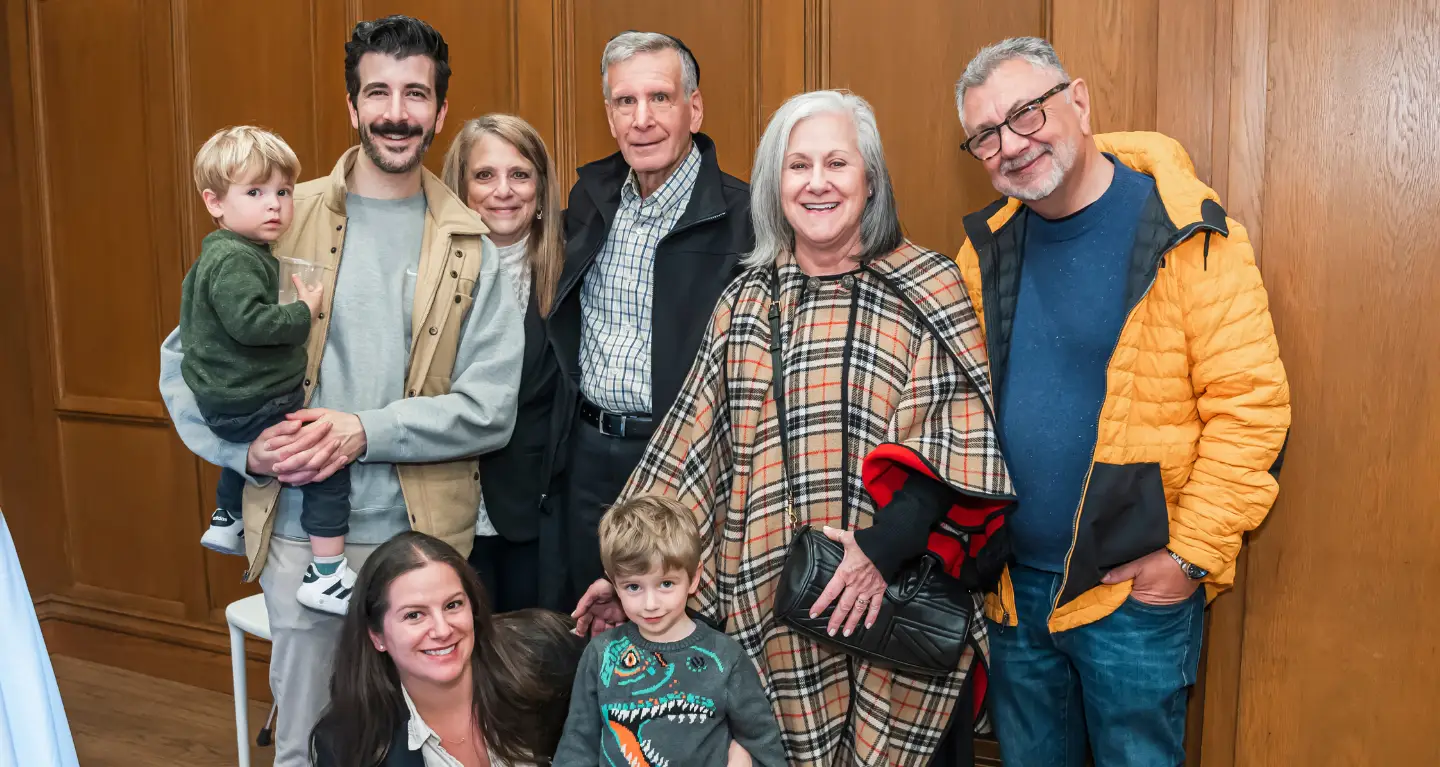 Seven adults and two young boys smile together in front of a wood-paneled wall at Temple Emanu-El in New York City.