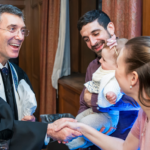 A rabbi wearing a tallit shakes hands with a woman holding a baby as the father looks on at Temple Emanu-El.