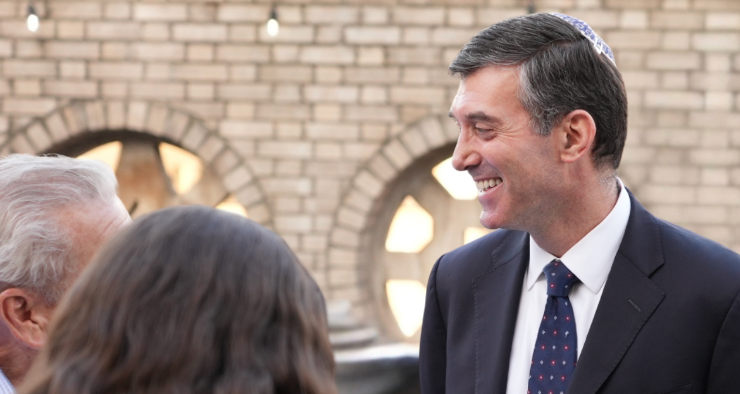 A man wearing a suit and yarmulke smiles while engaging in conversation with two people, standing in front of a brick wall featuring arched windows at Temple Emanu-El.