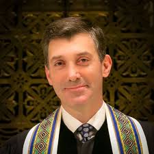 A rabbi in formal attire with a decorative tallit stands before an ornate, gold-colored background at Temple Emanu-El.
