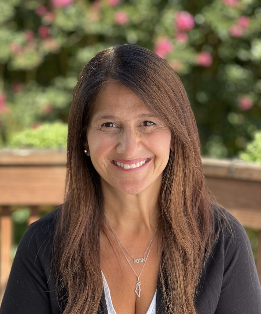 Smiling woman with long brown hair sits outdoors, with flowers and greenery softly blurred in the background.