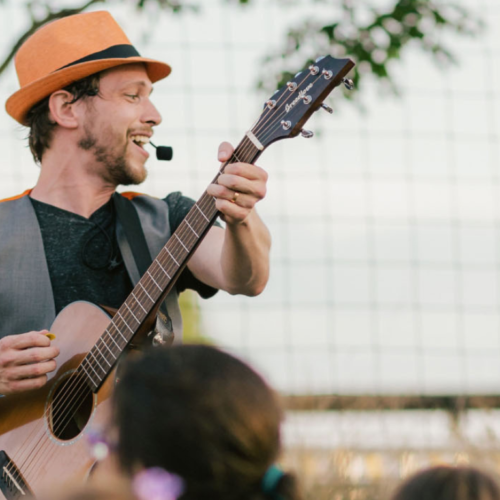 Man in orange hat playing guitar and singing outdoors at a Temple Emanu-El event, with attendees watching in the foreground.
