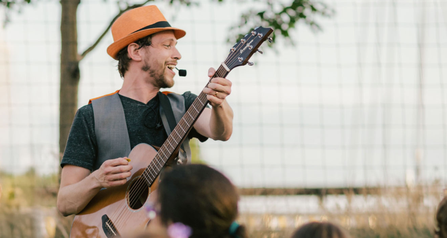 Man in orange hat playing guitar and singing outdoors at a Temple Emanu-El event, with attendees watching in the foreground.