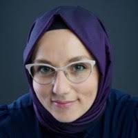 A woman wearing glasses and a deep purple headscarf smiles warmly at the camera, set against a dark background.