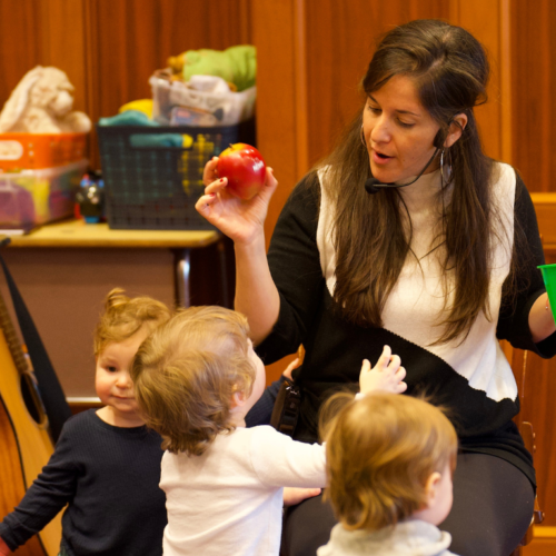 A woman teaches young children at Temple Emanu-El, holding a red apple and green basket, with toys and guitar in the background.