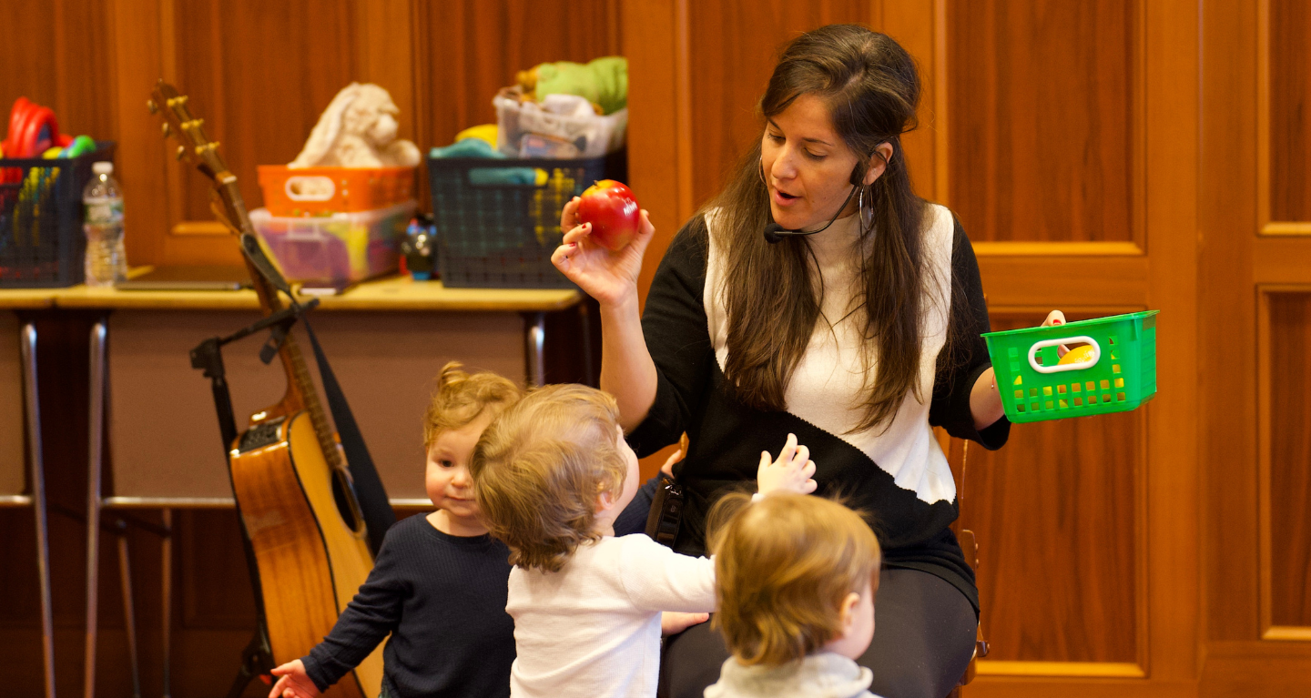 A woman teaches young children at Temple Emanu-El, holding a red apple and green basket, with toys and guitar in the background.