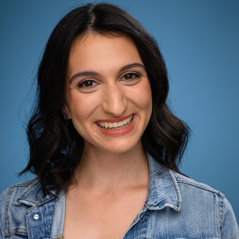 Smiling woman with dark hair in a denim jacket stands before a blue backdrop.