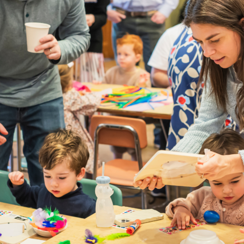 Adults and children creating crafts together at a busy table in a colorful, lively Temple Emanu-El classroom setting.
