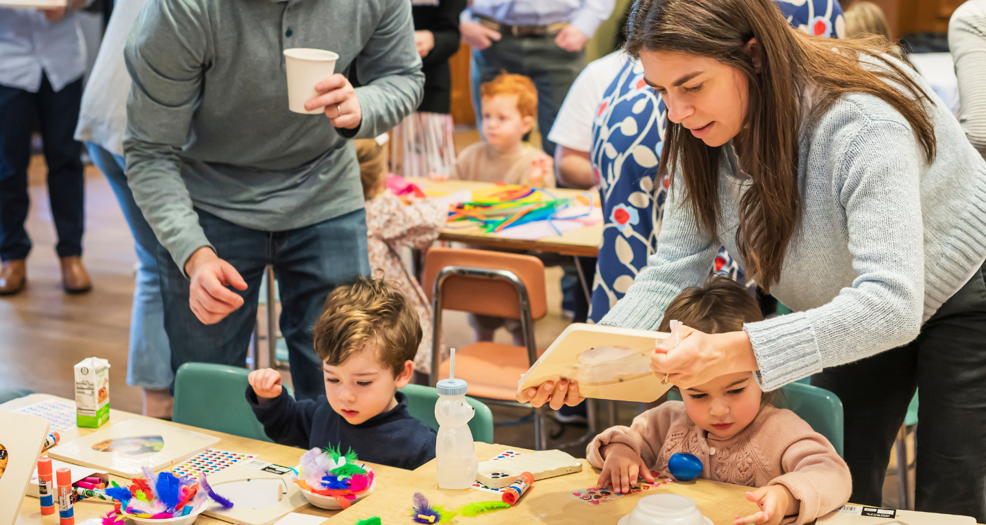 Adults and children creating crafts together at a busy table in a colorful, lively Temple Emanu-El classroom setting.