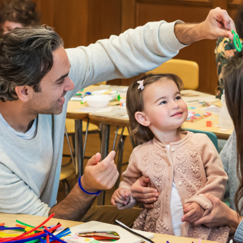 A family helps their toddler with a craft project at a colorful, busy classroom table in Temple Emanu-El’s religious school.