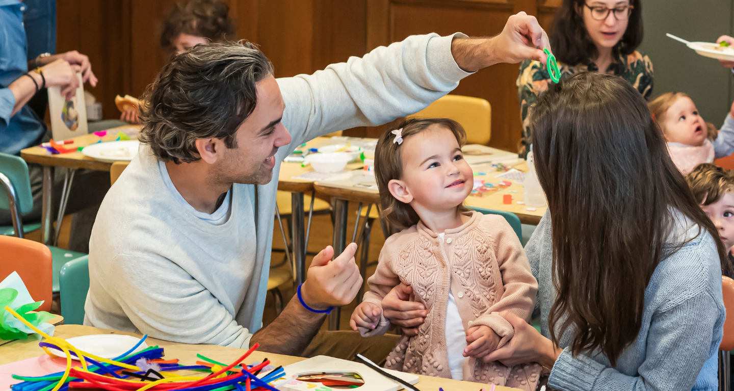 A family helps their toddler with a craft project at a colorful, busy classroom table in Temple Emanu-El’s religious school.