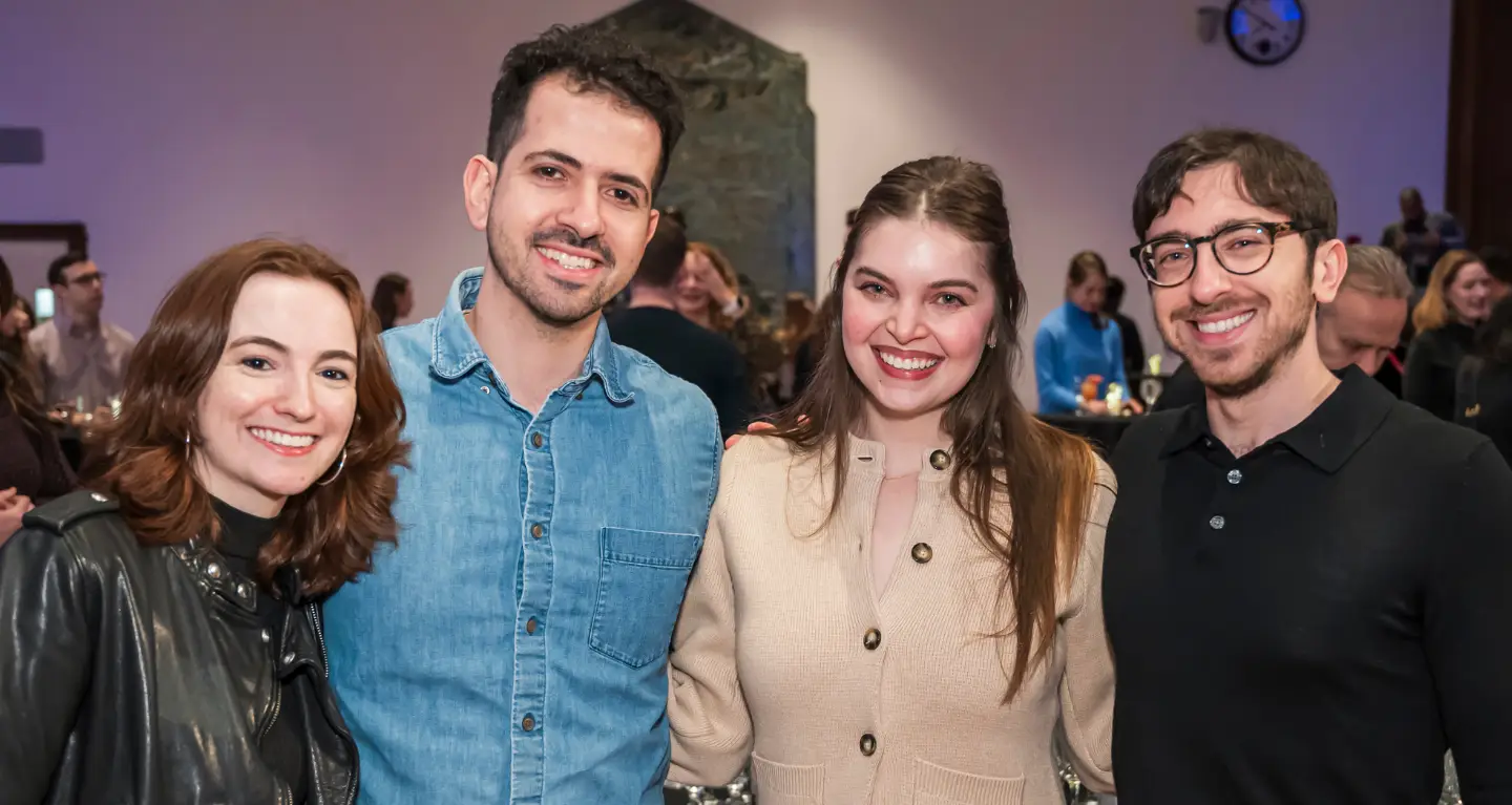 Four congregants smiling and posing together at a Temple Emanu-El community gathering, with others in the background.