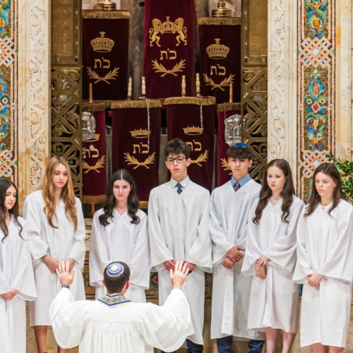A group of teens in white robes stand before a rabbi at Temple Emanu-El, surrounded by elegant floral arrangements.