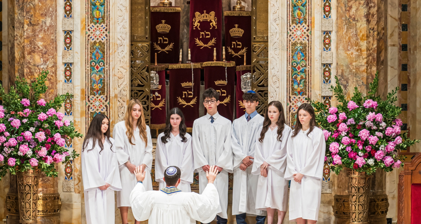 A group of teens in white robes stand before a rabbi at Temple Emanu-El, surrounded by elegant floral arrangements.