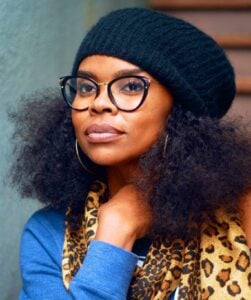 Woman with curly hair, glasses, a black beanie, and a leopard print scarf, smiling gently at the camera inside Temple Emanu-El.