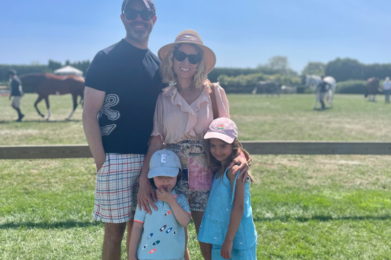 A family of four smiles together in a sunny field with horses, all dressed in light summer clothing and sun hats.