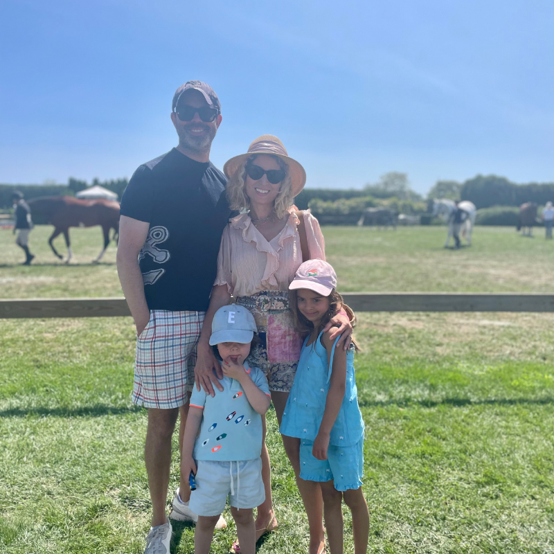 A family of four smiles together in a sunny field with horses, all dressed in light summer clothing and sun hats.