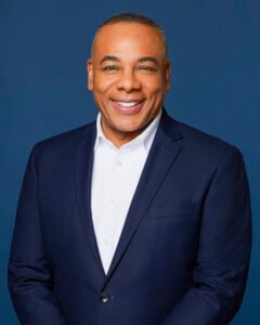 Smiling man in a navy suit and white shirt stands against a plain blue background at Temple Emanu-El in New York City.