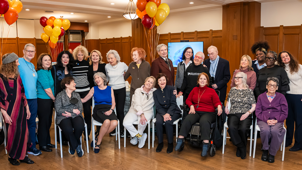 A diverse group of congregants gather and smile together inside Temple Emanu-El with festive balloons in the background.