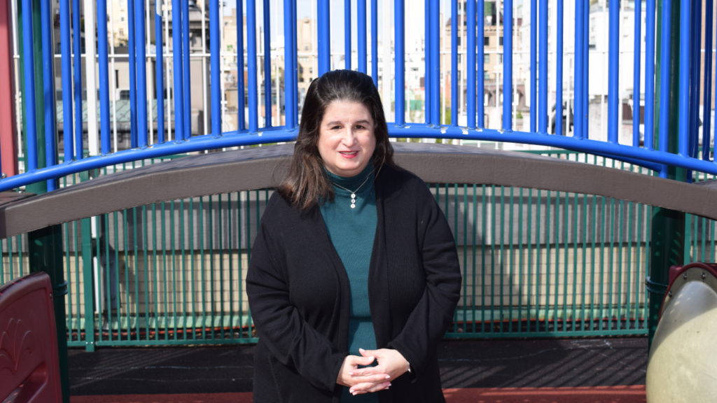 Woman with dark hair in a black coat smiles at the camera while standing outdoors near blue playground bars at Temple Emanu-El.