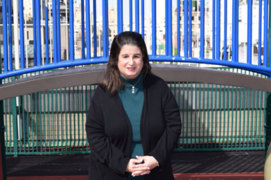 Woman with dark hair in a black coat smiles at the camera while standing outdoors near blue playground bars at Temple Emanu-El.
