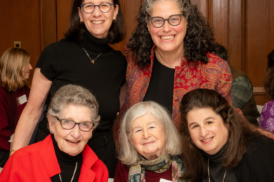 Five women smile together at a Temple Emanu-El event, wearing name tags and dressed in dark and red attire indoors.