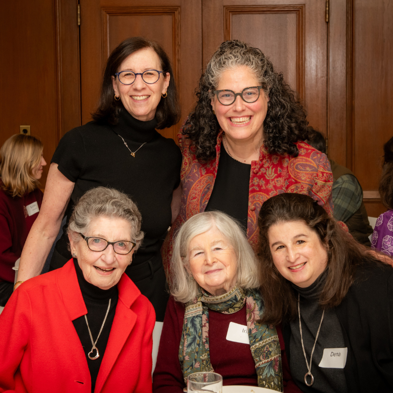Five women smile together at a Temple Emanu-El event, wearing name tags and dressed in dark and red attire indoors.