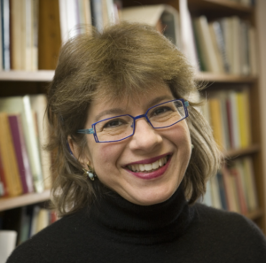Woman with brown hair, blue glasses, and black sweater smiling warmly in front of a bookshelf at Temple Emanu-El in New York City.