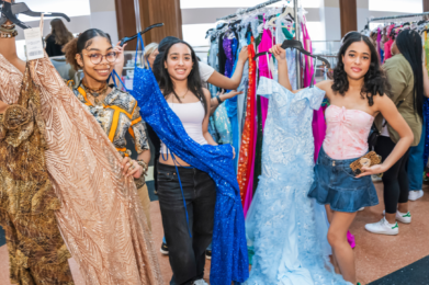 Three young women smiling and holding up vibrant dresses in a shop, with racks of additional dresses visible behind them.