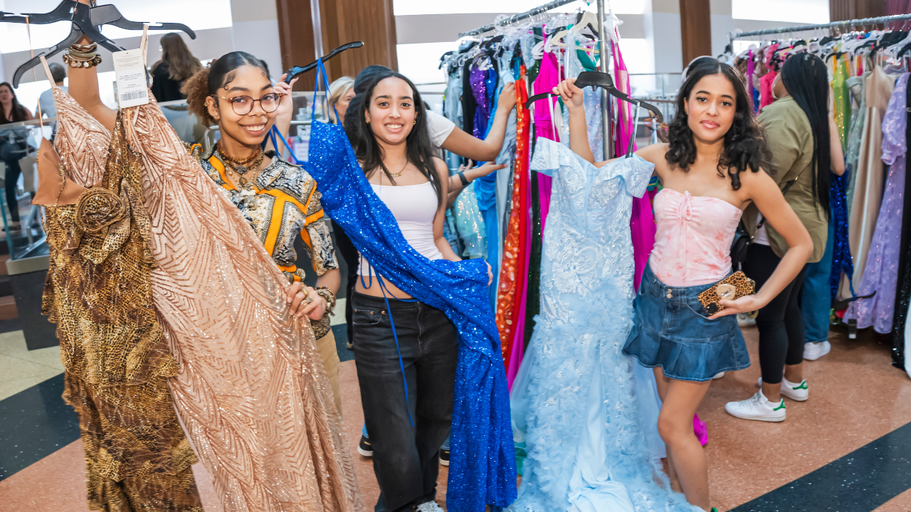 Three young women smiling and holding up vibrant dresses in a shop, with racks of additional dresses visible behind them.