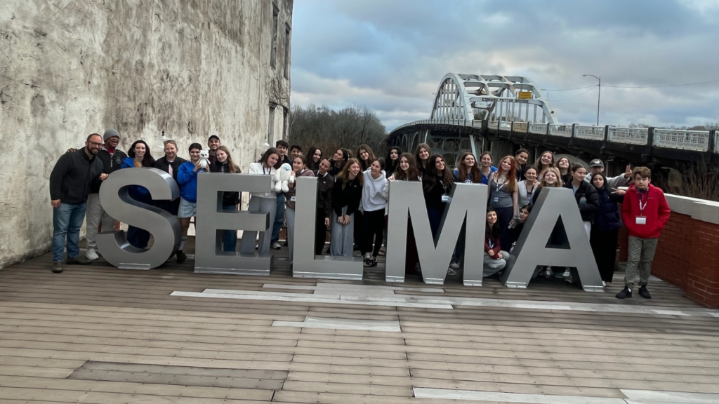 Members of Temple Emanu-El stand behind large SELMA letters by a bridge under a cloudy sky, honoring civil rights history.