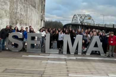 Members of Temple Emanu-El stand behind large SELMA letters by a bridge under a cloudy sky, honoring civil rights history.