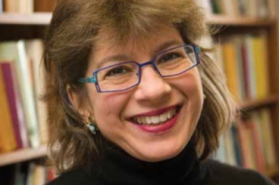 A smiling individual with glasses and brown hair, dressed in a black top, stands before bookshelves.