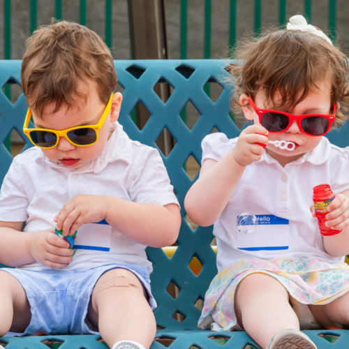 Two young children wearing sunglasses sit together on a bench; one blows bubbles while the other examines a bubble wand.